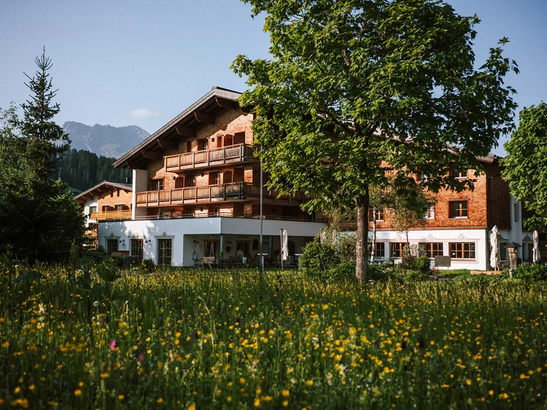 Aurora, Ihr Hotel in Lech am Arlberg Haus mit Holzfassade neben Bäumen und Wiese mit gelben Blumen unter blauem Himmel