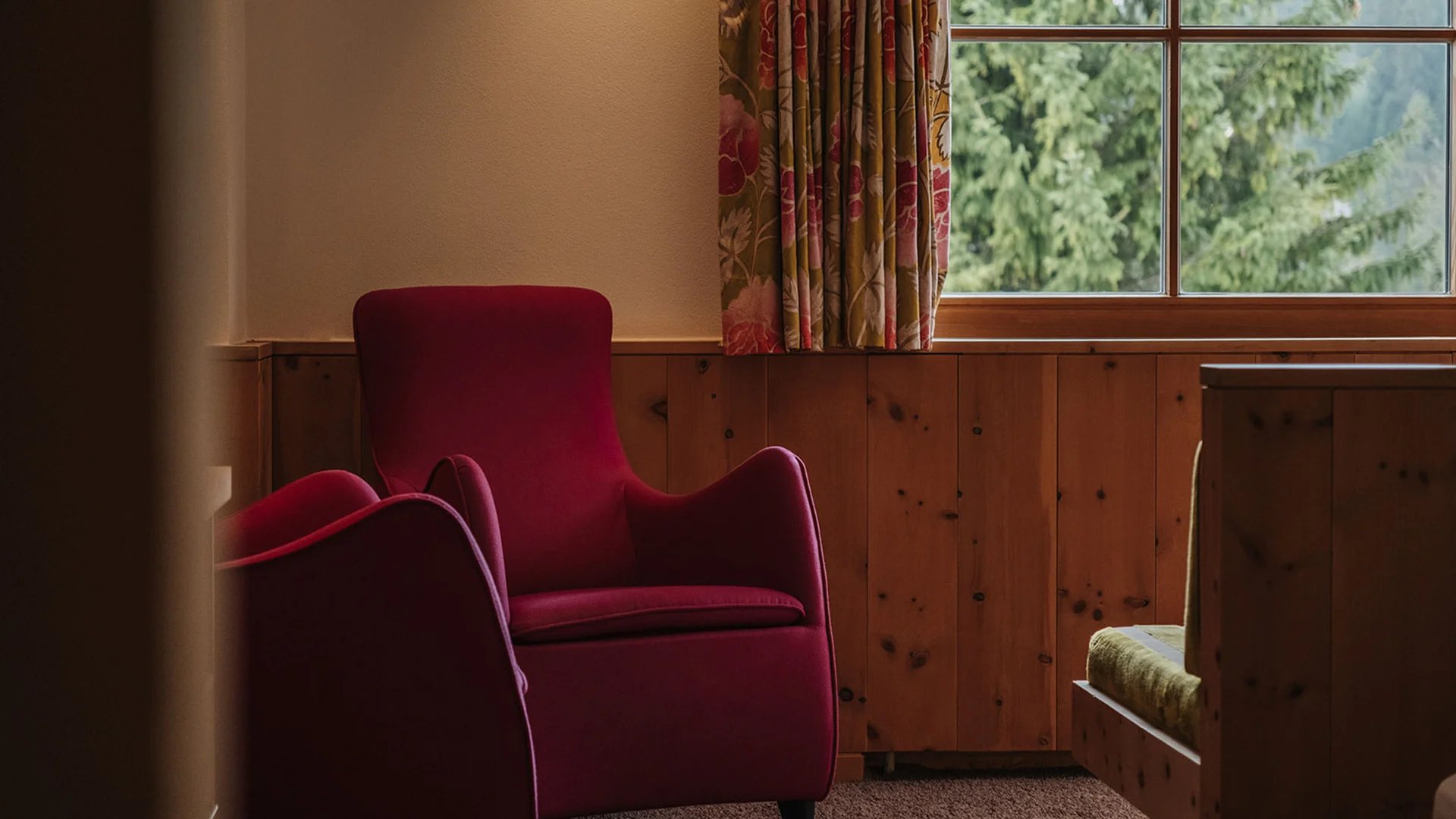 Aurora, Ihr Hotel in Lech am Arlberg Roter Sessel vor einem Fenster mit Blick auf grüne Bäume in einem Raum mit Holzvertäfelung