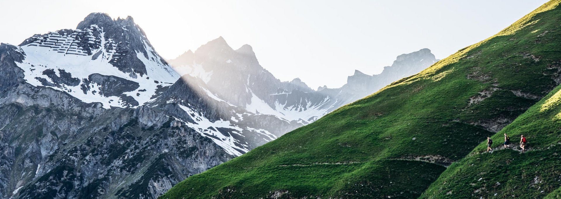 Drei Personen wandern an einem schmalen Pfad entlang mit Ausblick auf die schroffen Gipfel. 