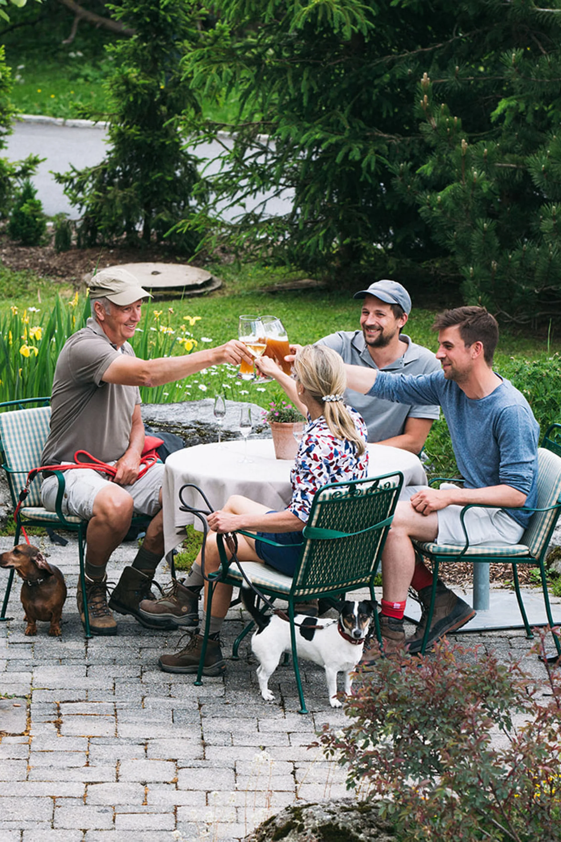 Aurora, Ihr Hotel in Lech am Arlberg Vier Personen stoßen mit Getränken im Garten an, zwei kleine Hunde sind dabei